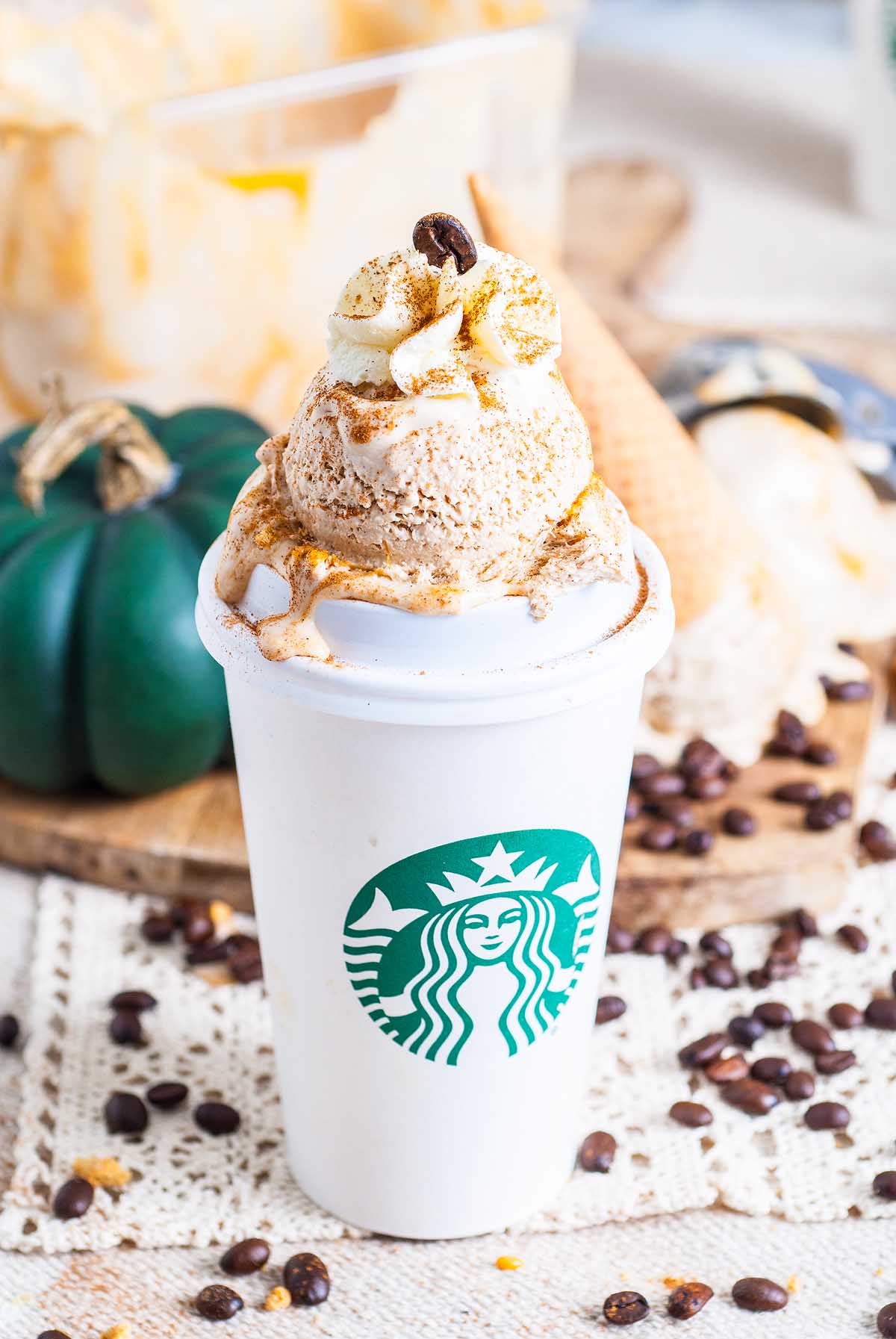 A Starbucks cup filled with no churn pumpkin spice latte ice cream, topped with whipped cream and coffee beans, with scattered coffee beans around.