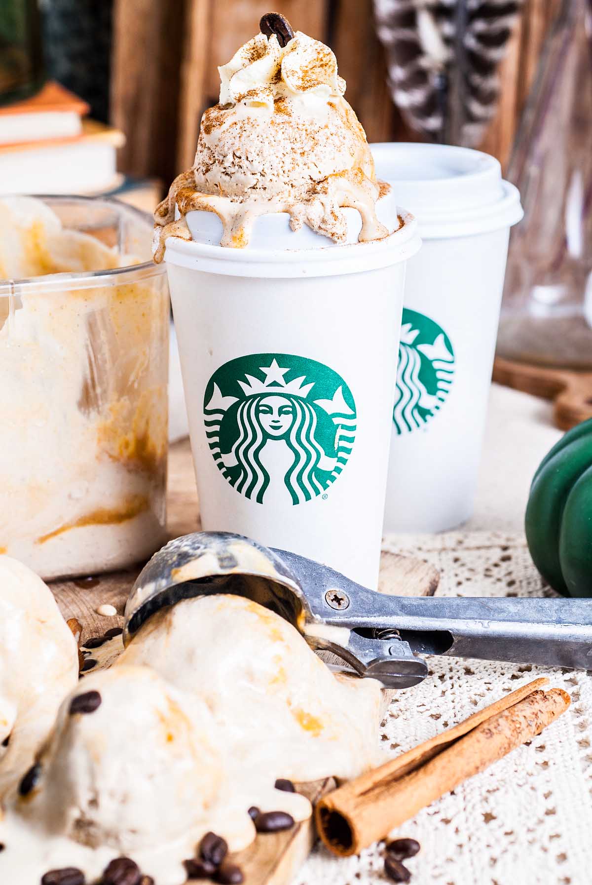 Starbucks cup filled with no churn pumpkin spice latte ice cream and whipped cream, surrounded by ice cream scoops and coffee beans.