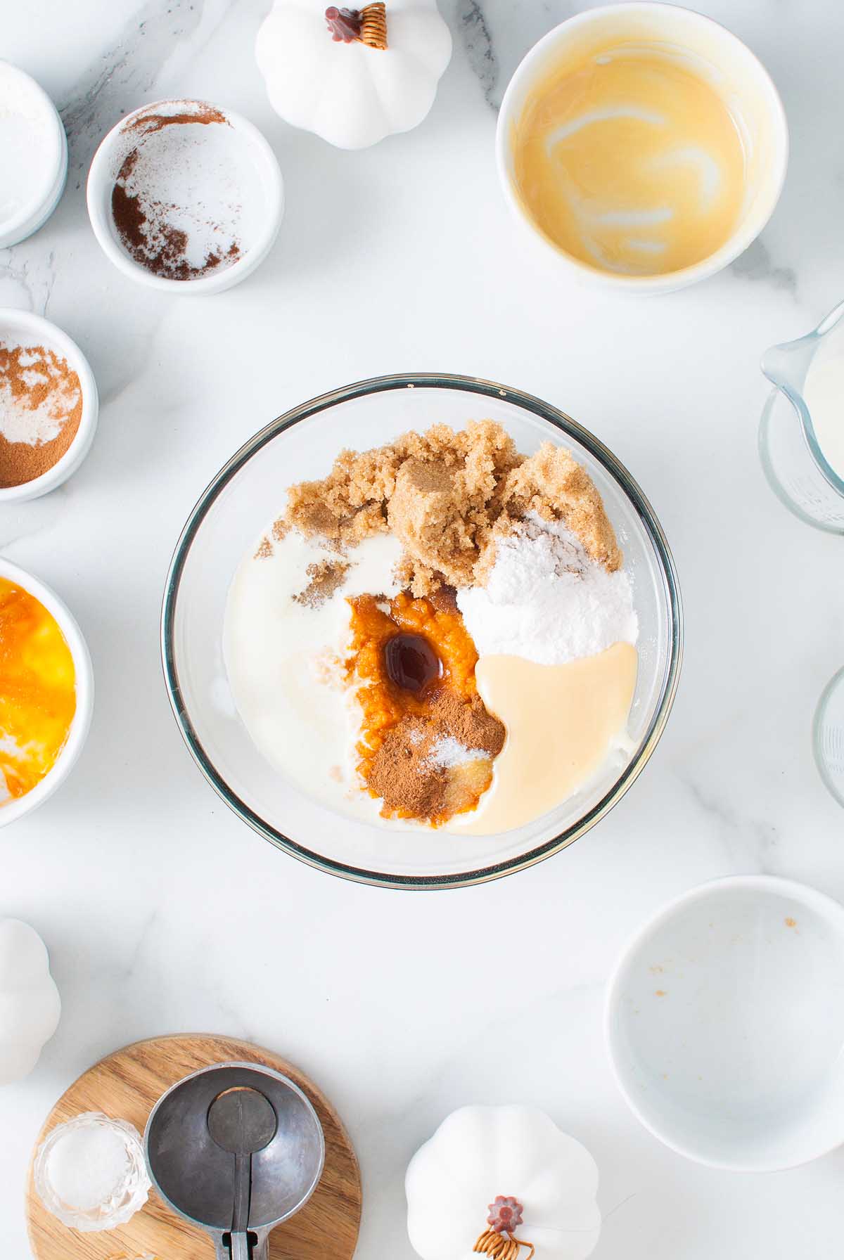 A bowl of baking ingredients on a white surface, surrounded by spices, eggs, and small white pumpkins—perfect for creating no churn pumpkin spice latte ice cream.