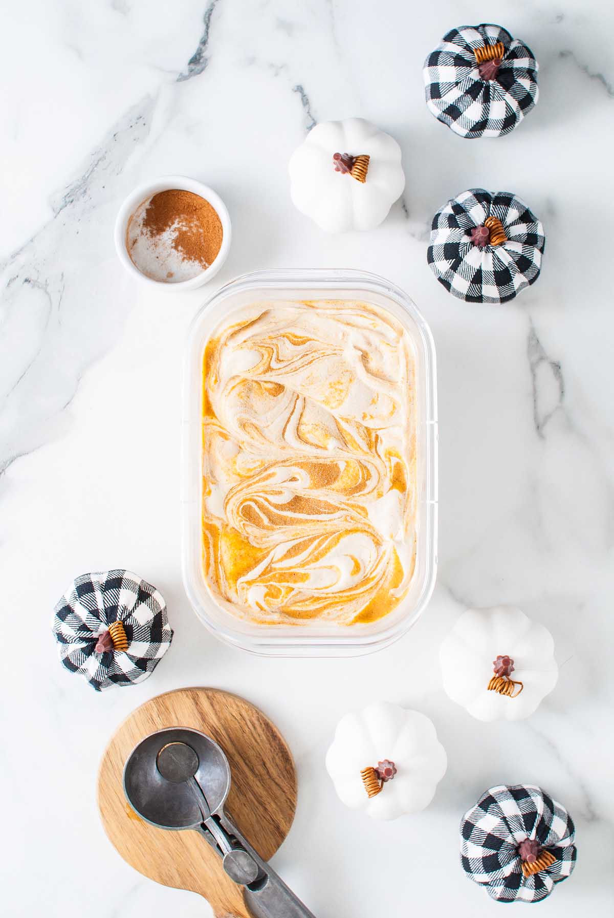 No churn pumpkin spice latte ice cream swirled in a container, surrounded by small white pumpkins, black plaid pumpkins, and a scoop on marble.