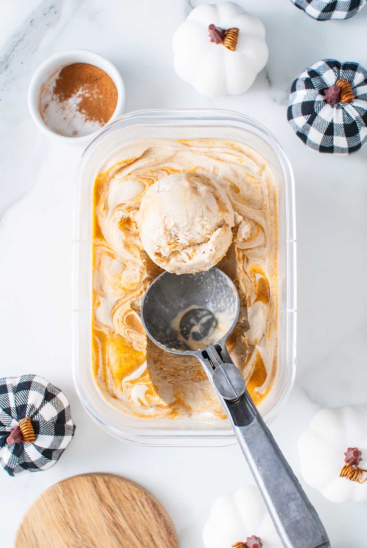 A tub of no churn pumpkin spice latte ice cream with a scoop, surrounded by small pumpkins and a bowl of cinnamon.