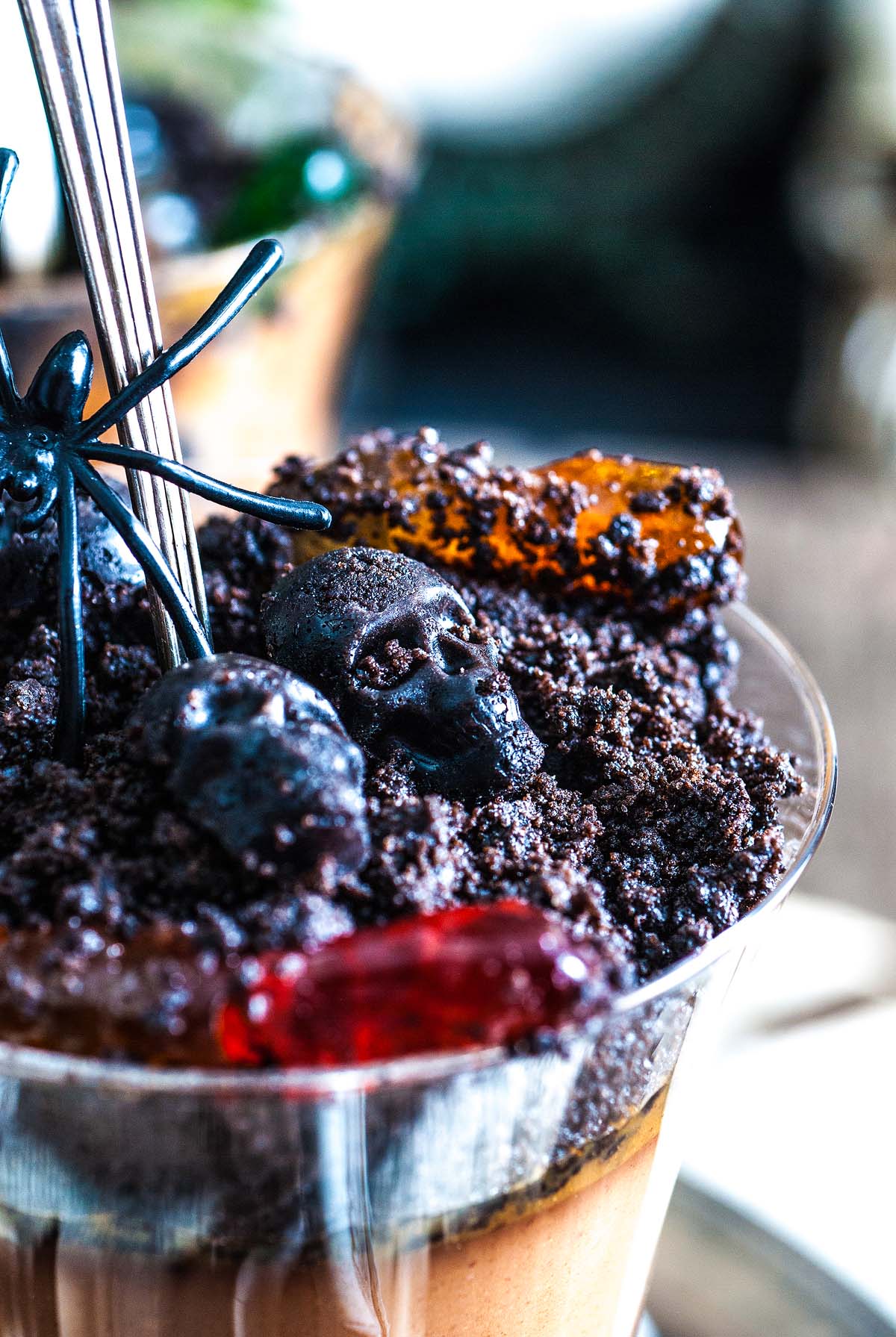 A close-up of a Boozy Halloween Pot de Creme Dessert, topped with cookie crumbs, gummy worms, and candy skulls for the perfect spooky Halloween dessert.