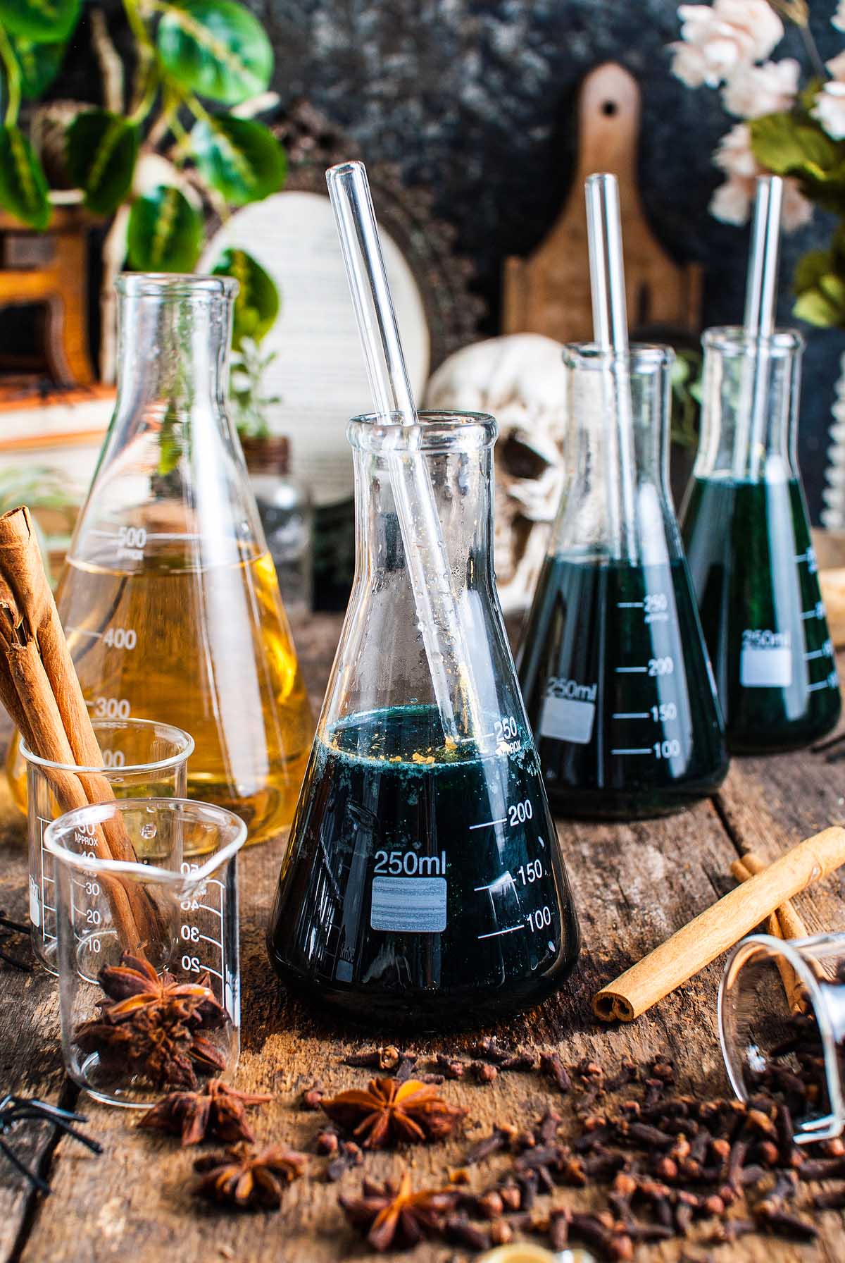 Assorted glass beakers with shimmering Halloween apple cider punch, dark and amber drinks, cinnamon sticks, and star anise on a rustic wooden table.
