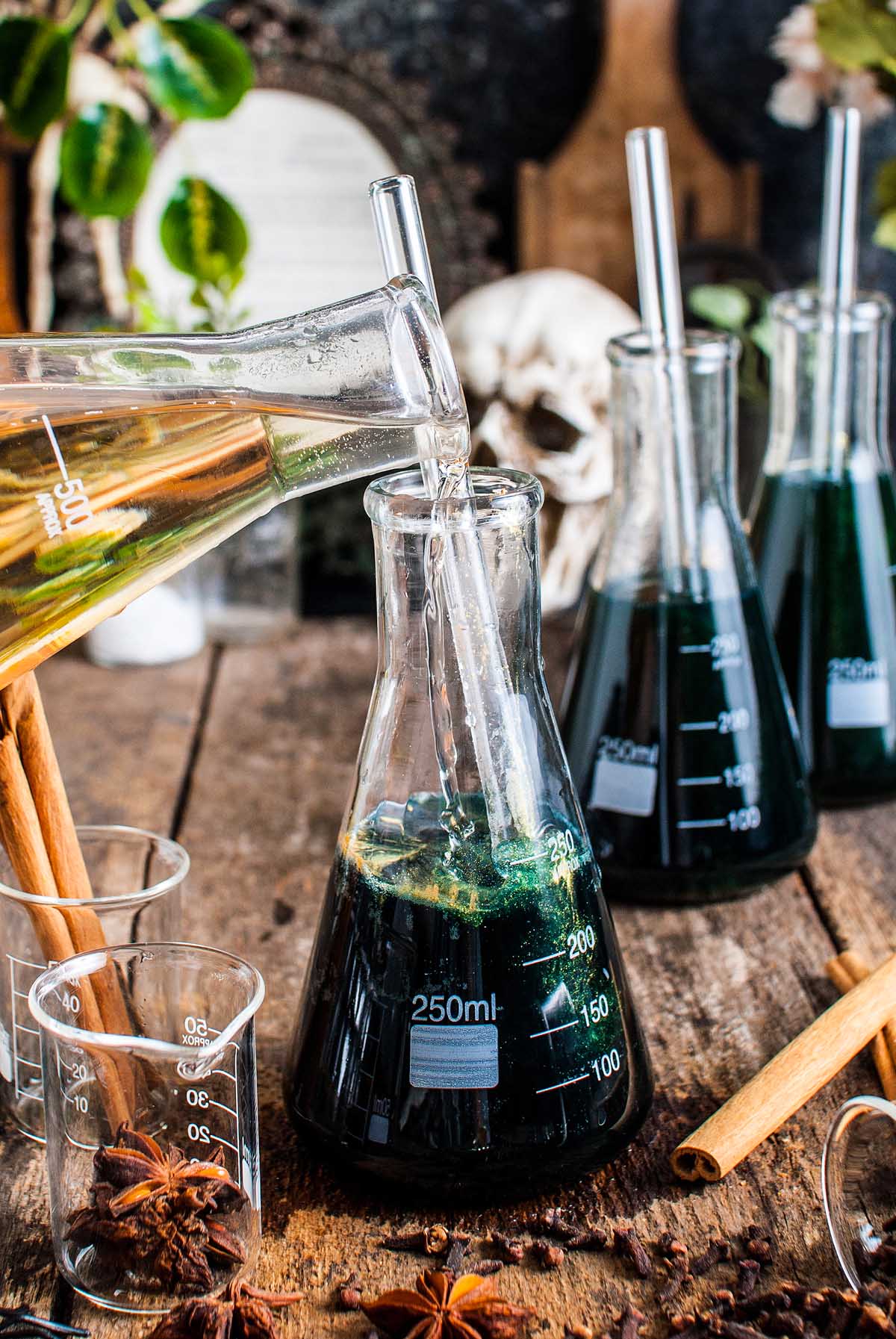 A beaker pours shimmering Halloween apple cider punch into a flask with dark green fluid, set on a rustic table adorned with lab glassware and spices.