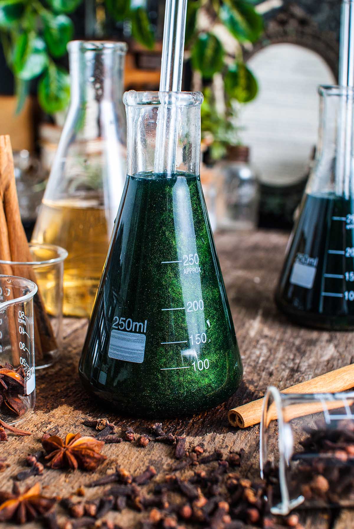 A flask filled with dark green Shimmering Halloween apple cider punch sits on a wooden table, surrounded by spices and other laboratory glassware.