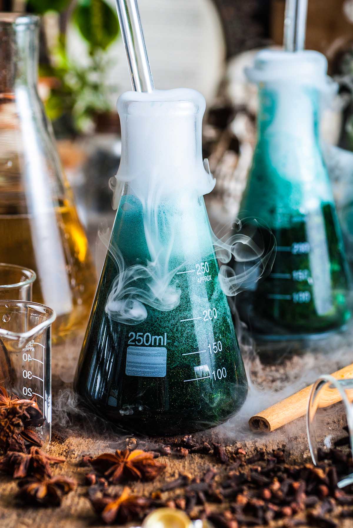 A steaming green liquid, like a shimmering Halloween apple cider punch, sits in a 250ml flask emitting vapor on a rustic table surrounded by spices and other glassware.