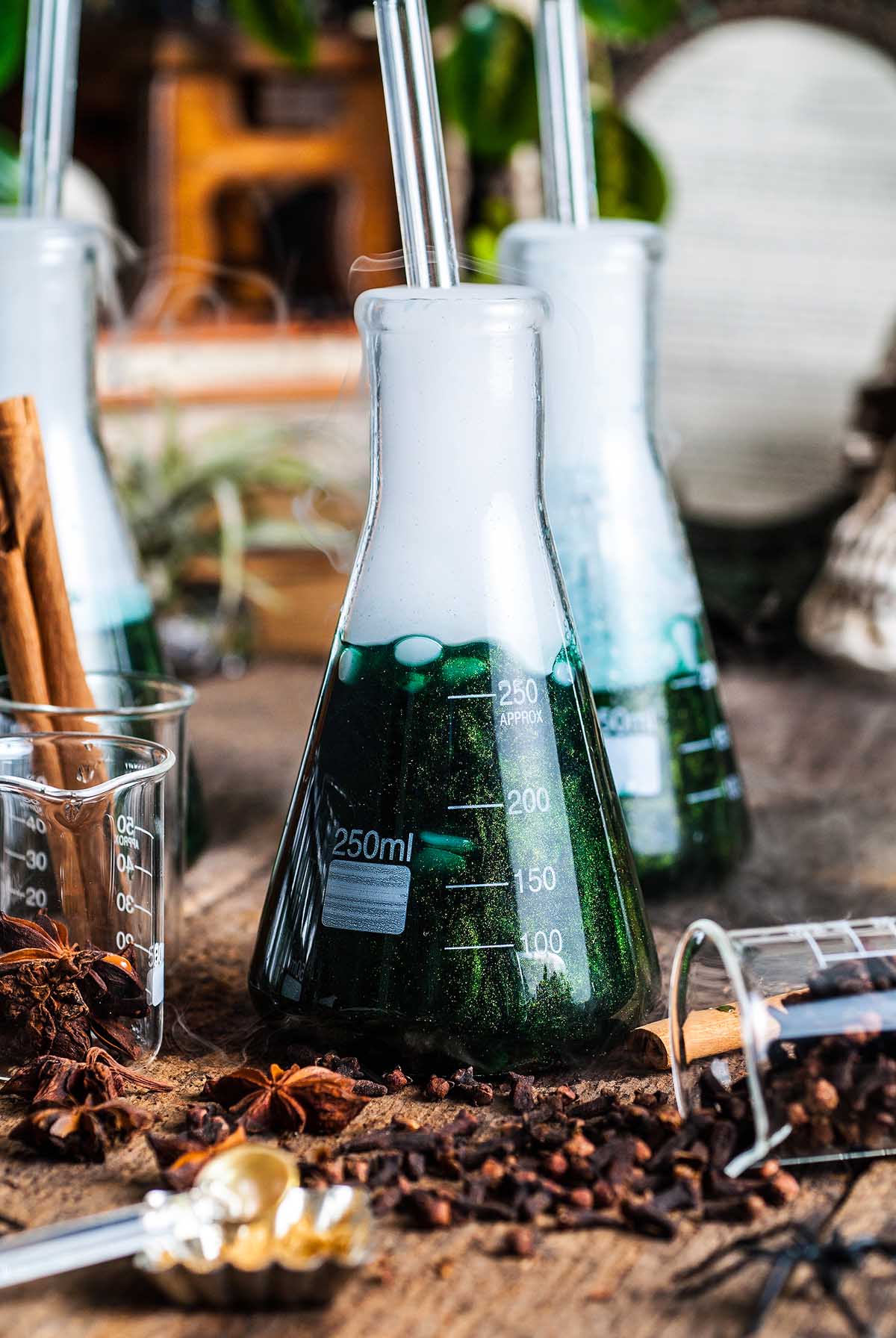A green fizzy Shimmering Halloween apple cider punch bubbles in a 250ml lab flask with a straw, surrounded by spices and beakers on a wooden surface.