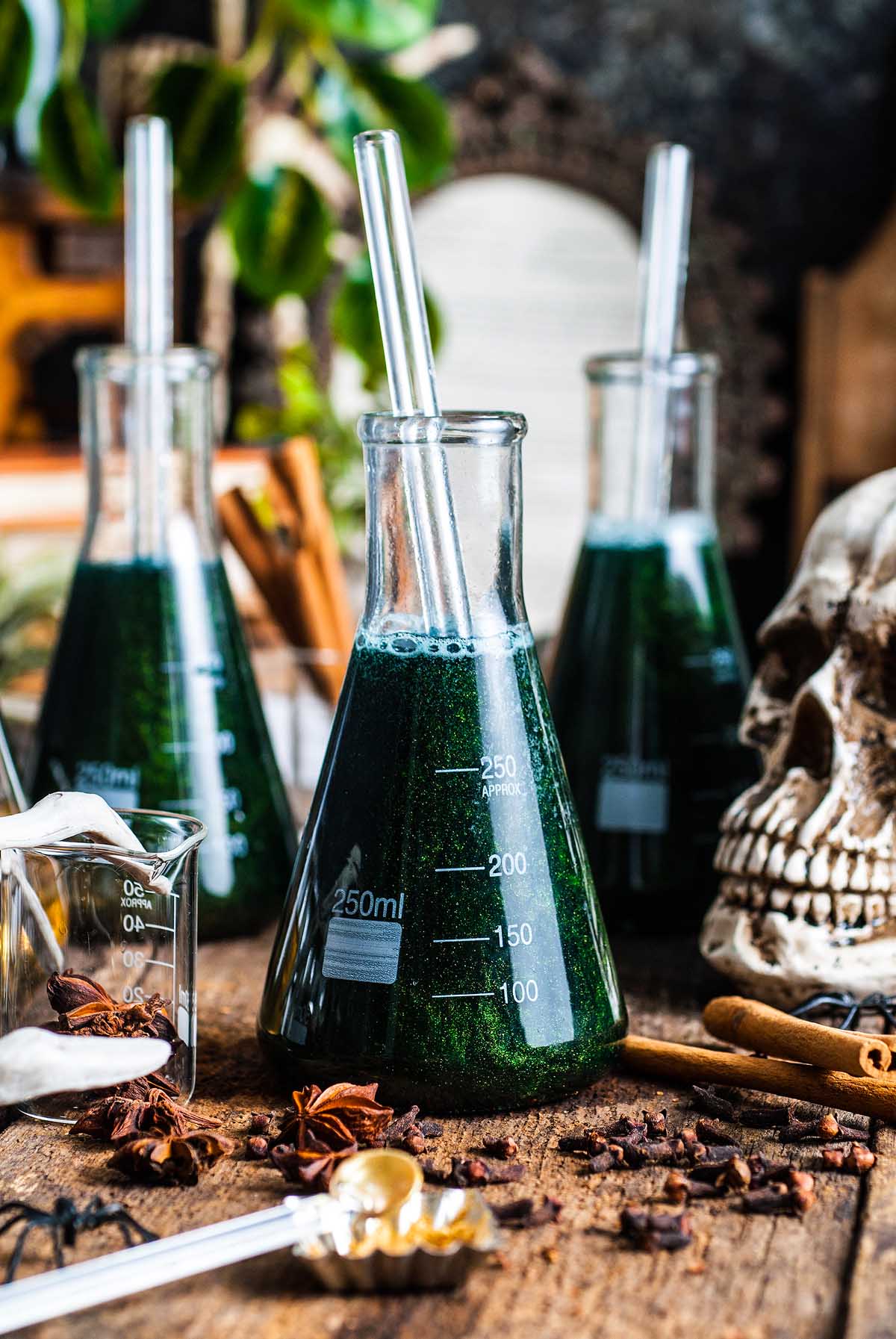 A green shimmering Halloween apple cider punch sits in a science beaker with a straw, surrounded by spices and a skull on a rustic table.
