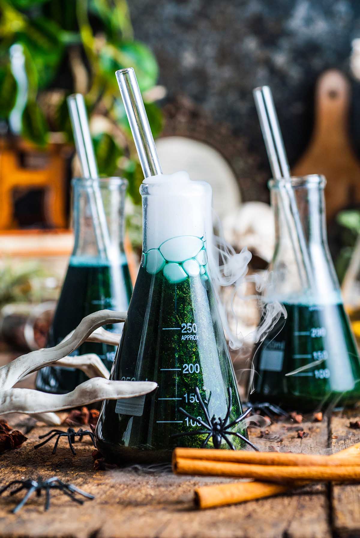 Three beakers filled with bubbling, green Shimmering Halloween apple cider punch, with straws and fake spiders on a rustic wooden table.
