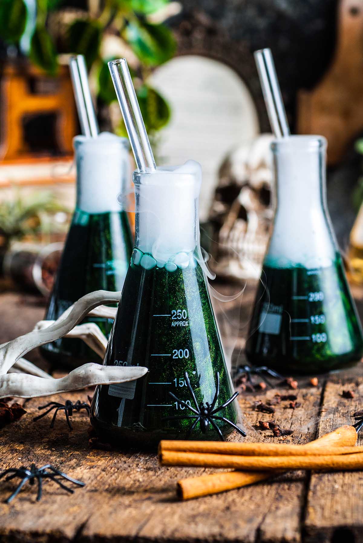 Three beakers with shimmering Halloween apple cider punch bubble with green drinks and straws, surrounded by fake spiders and spooky Halloween decorations.