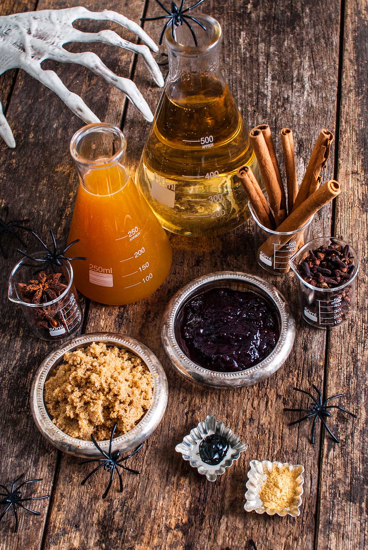 Shimmering Halloween apple cider punch ingredients in beakers on a wooden table, surrounded by spiders and a skeleton hand prop.
