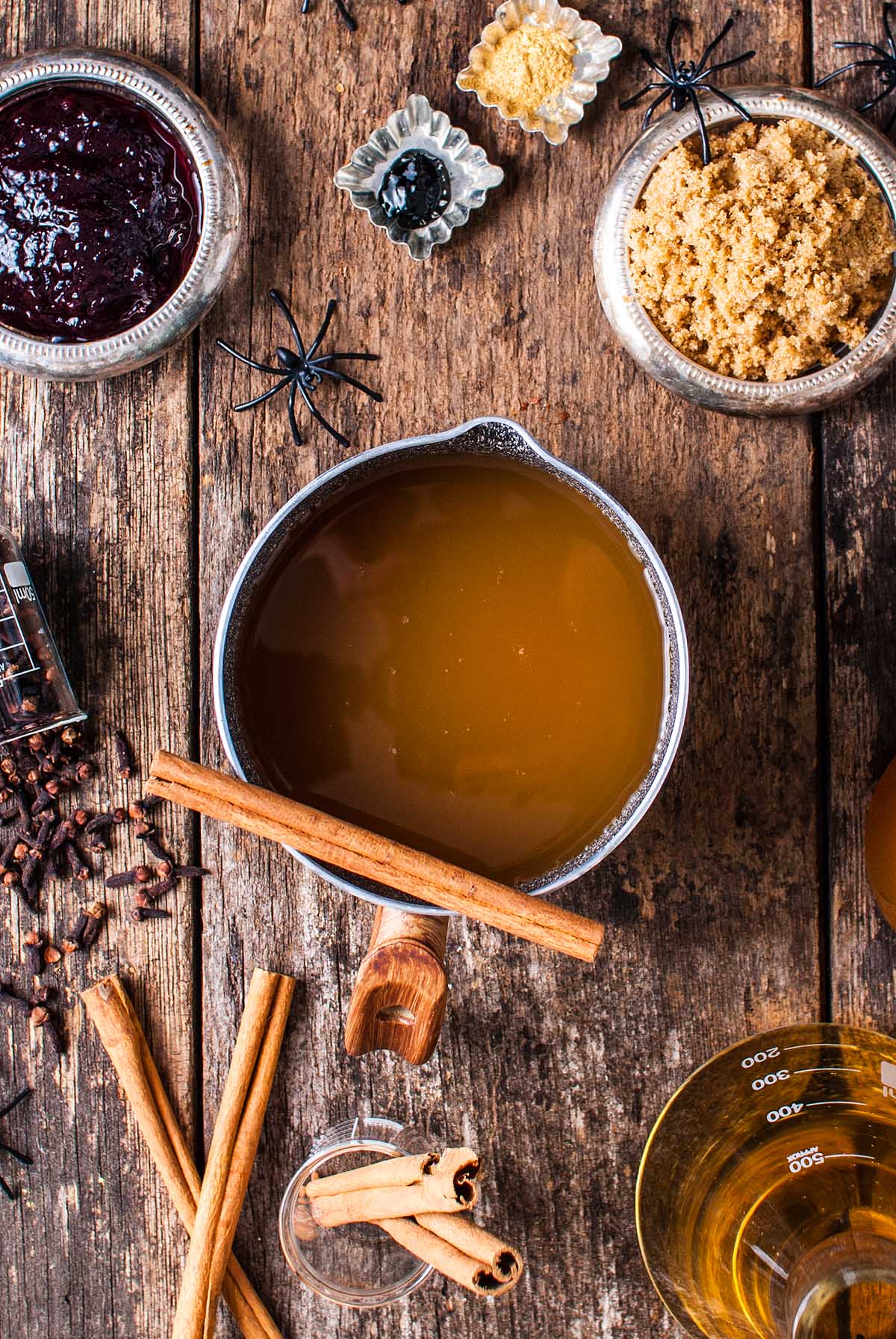 Overhead view of shimmering Halloween apple cider punch with cinnamon sticks, brown sugar, cloves, jam, and festive spider decorations.