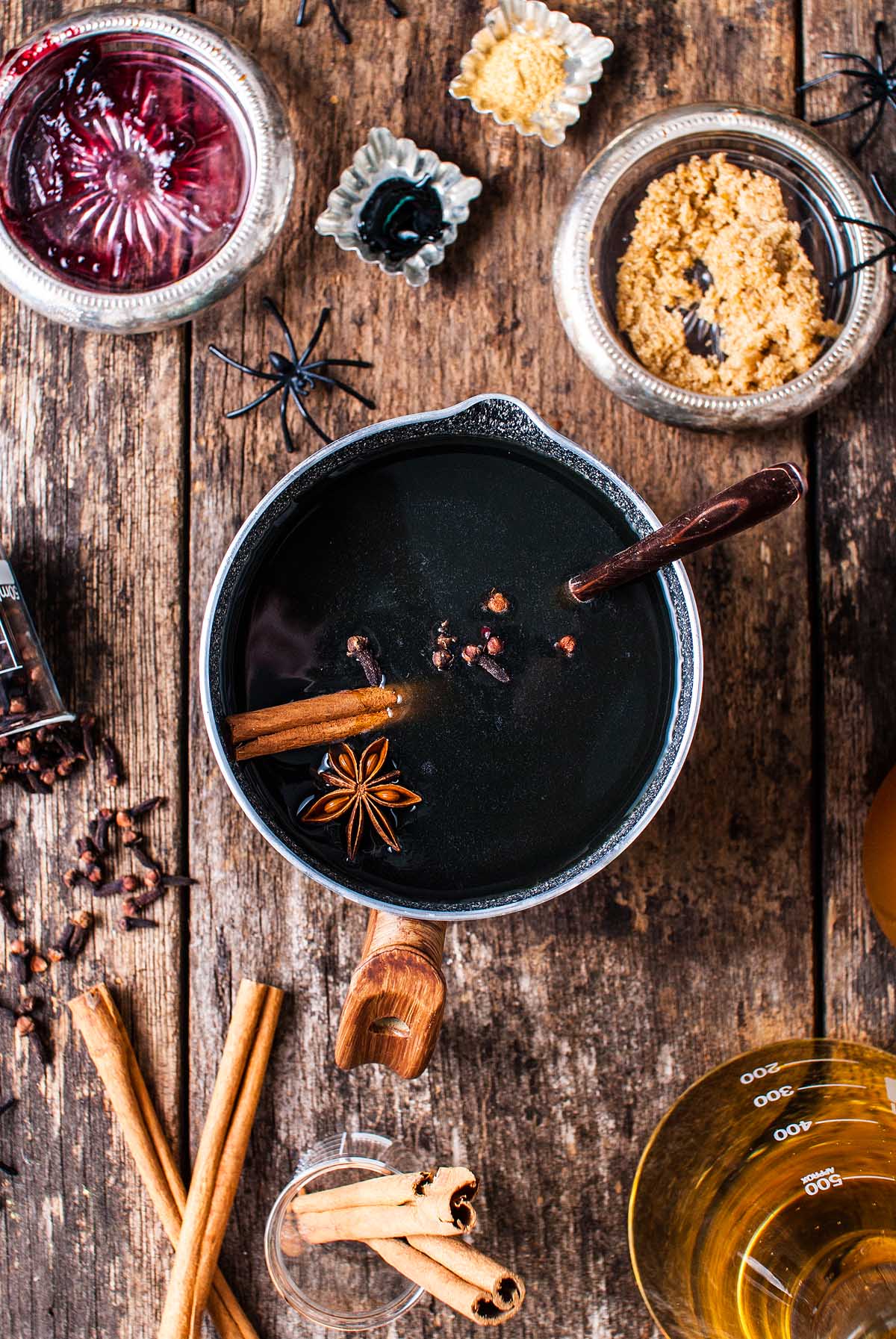 A pot of dark beverage with spices sits on a rustic table, surrounded by ingredients and fake spiders—perfect for serving as a shimmering Halloween apple cider punch.
