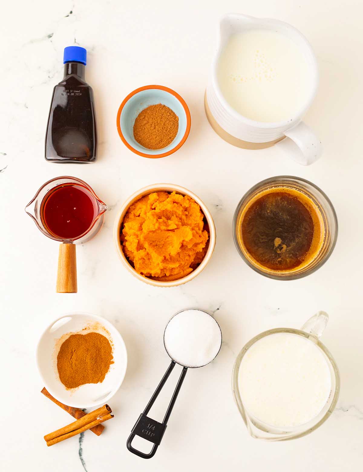 Ingredients for a slow cooker pumpkin spice latte arranged on a white surface, including puree, spices, milk, and coffee.