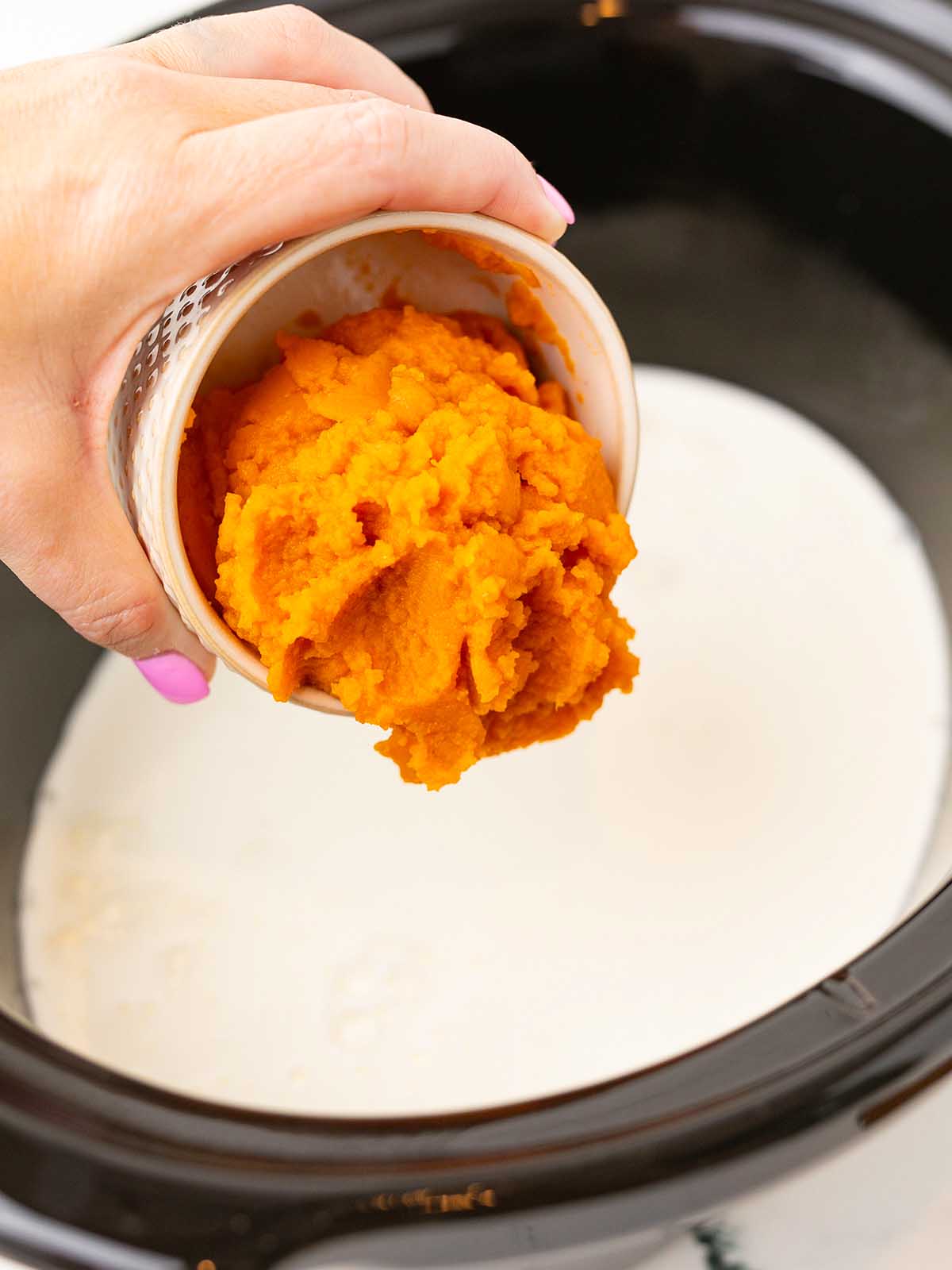 A hand pours canned pumpkin puree into a slow cooker with milk, preparing a cozy slow cooker pumpkin spice latte.