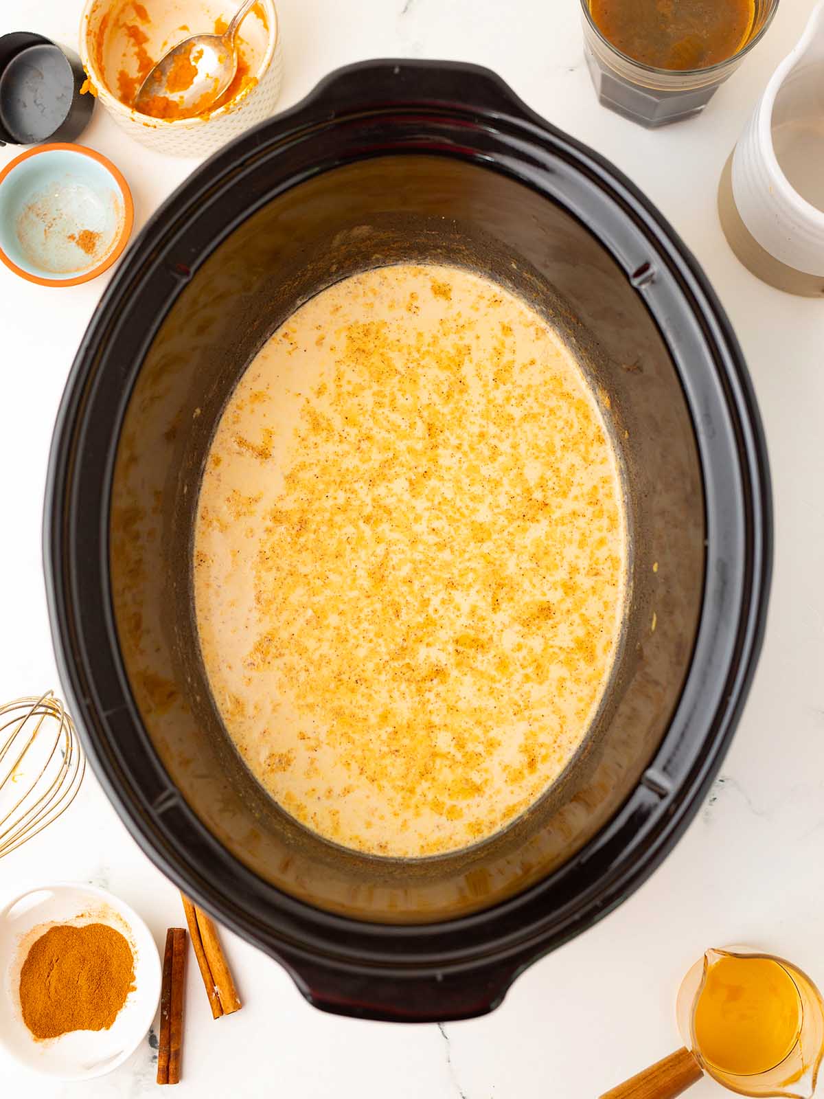 Slow cooker filled with a creamy, golden mixture for a cozy slow cooker pumpkin spice latte, surrounded by bowls of spices, cinnamon sticks, and milk.