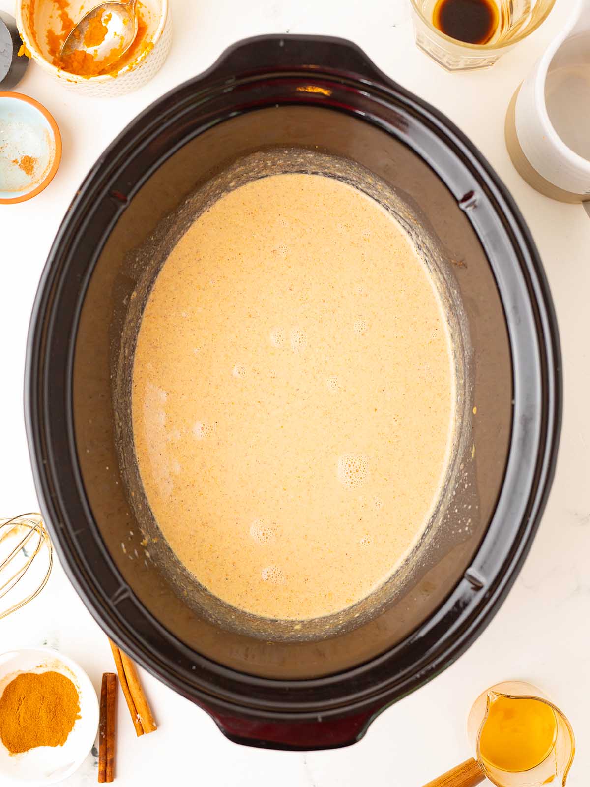 A slow cooker filled with creamy, tan slow cooker pumpkin spice latte, surrounded by spices and ingredients on a white surface.