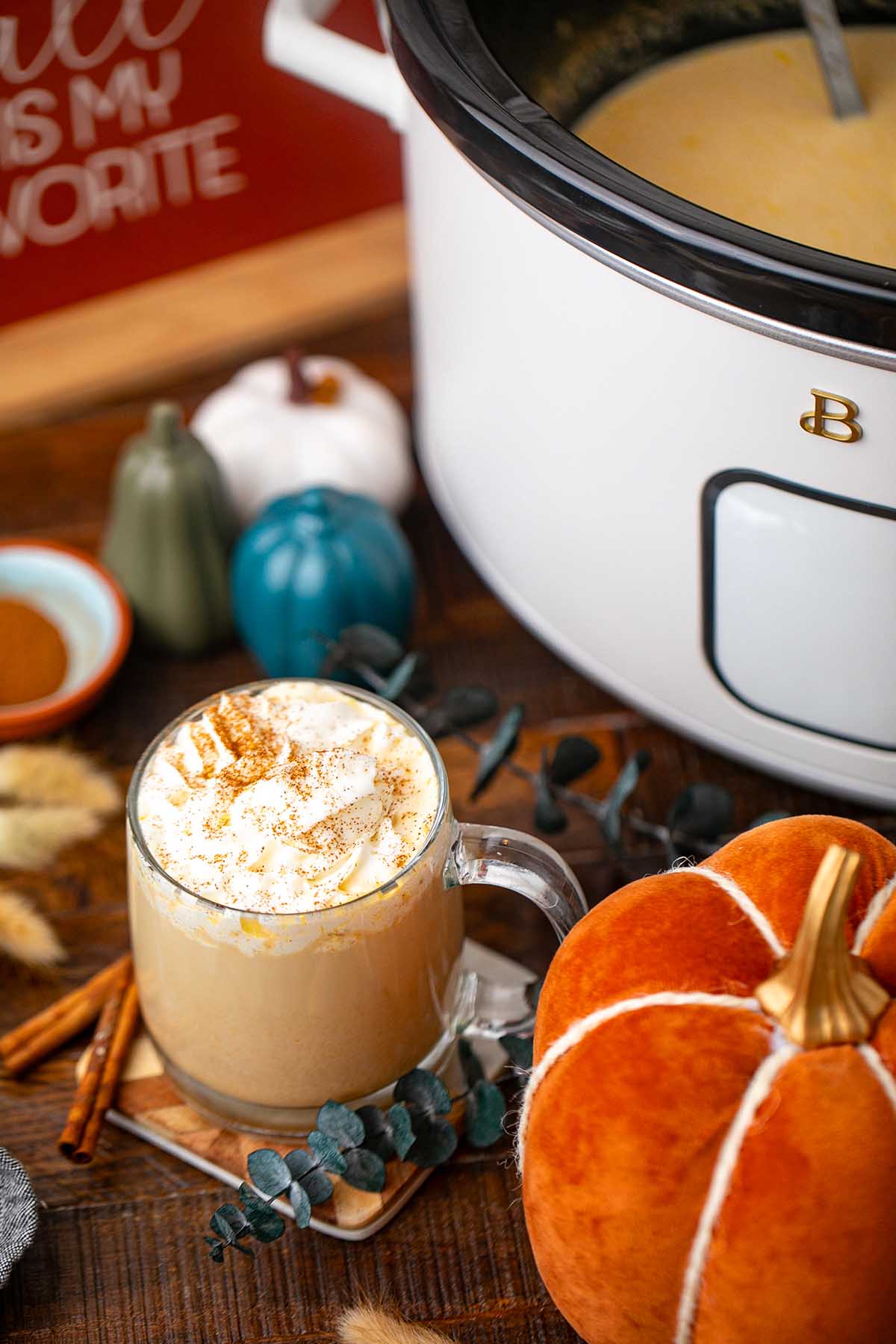 A mug of slow cooker pumpkin spice latte with whipped cream sits beside velvet pumpkins on a wooden table.