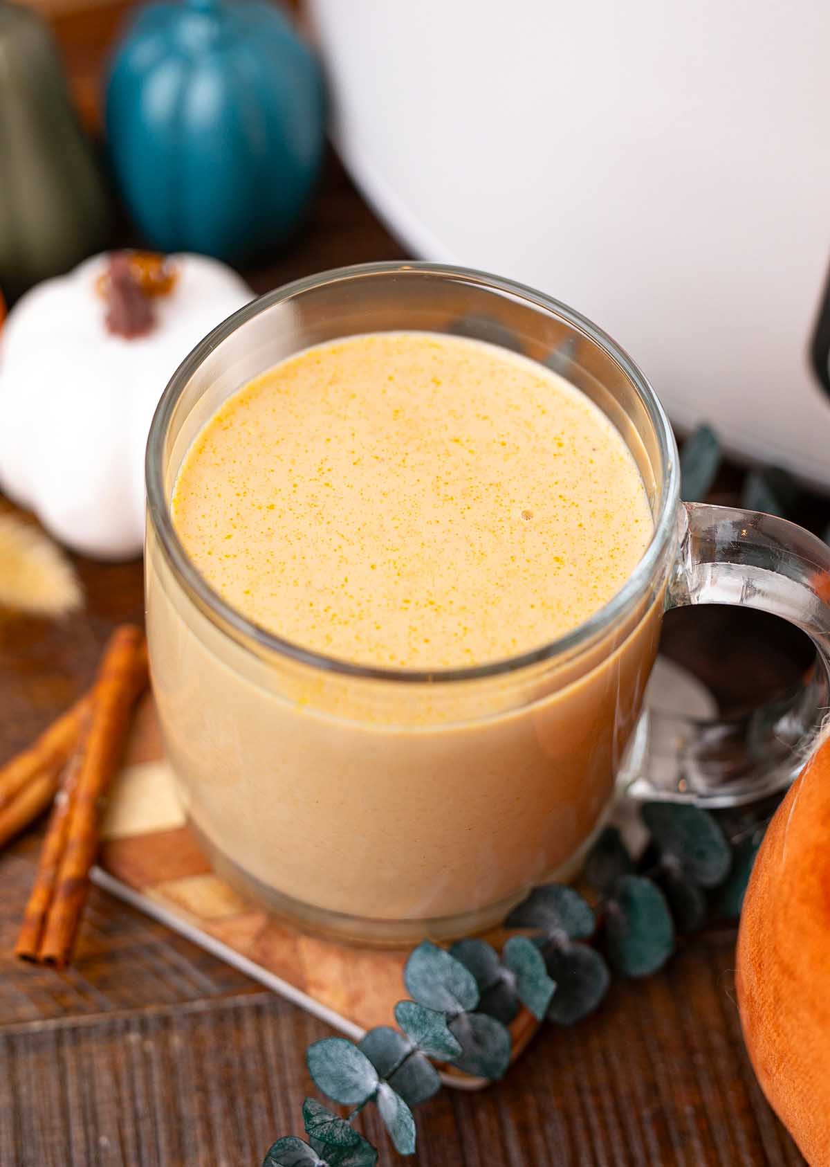 A clear mug of creamy slow cooker pumpkin spice latte sits on a wooden table with cinnamon sticks and fall decor nearby.