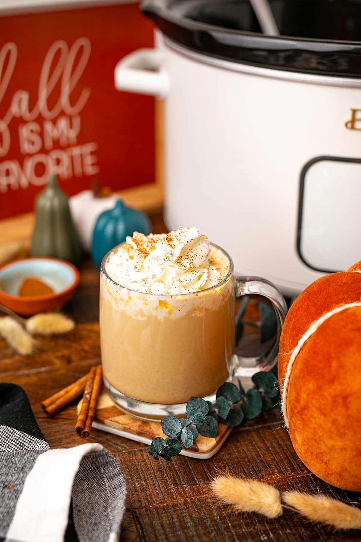 A glass mug of slow cooker pumpkin spice latte topped with whipped cream sits beside pumpkins and a slow cooker on a wooden table.