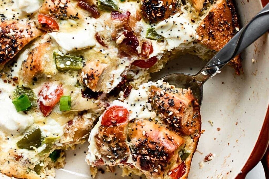 Everything bagel casserole with tomatoes and cream cheese, in a baking dish with a spoon.