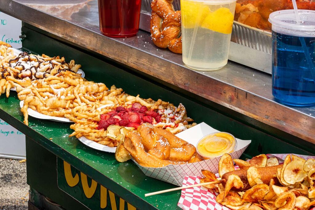 Delicious funnel cakes, a pretzel with mustard, and a corndog with potato chips stand on display outside of a festival food truck..