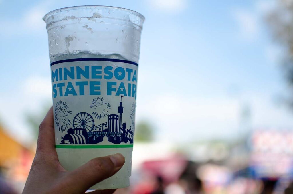 A hand holds up a Minnesota State Fair cup with a blurred background. The Minnesota State Fair runs for 12 days near the end of summer.