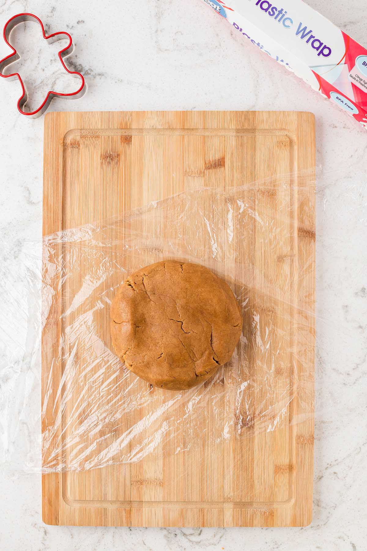 Ball of brown dough wrapped in plastic on a wooden board, with a gingerbread cutter and plastic wrap box nearby—perfect for making Easy Gingerbread Man Cookies.