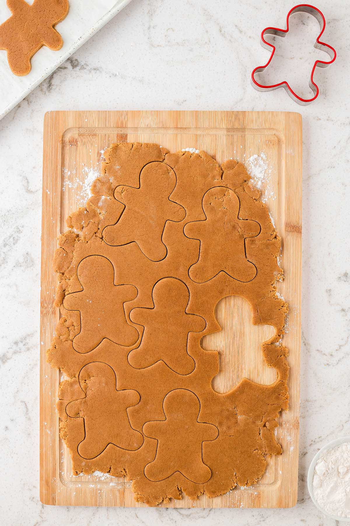 Rolled out Easy Gingerbread Man Cookies dough on a wooden board with gingerbread men shapes cut out and a red cookie cutter nearby.