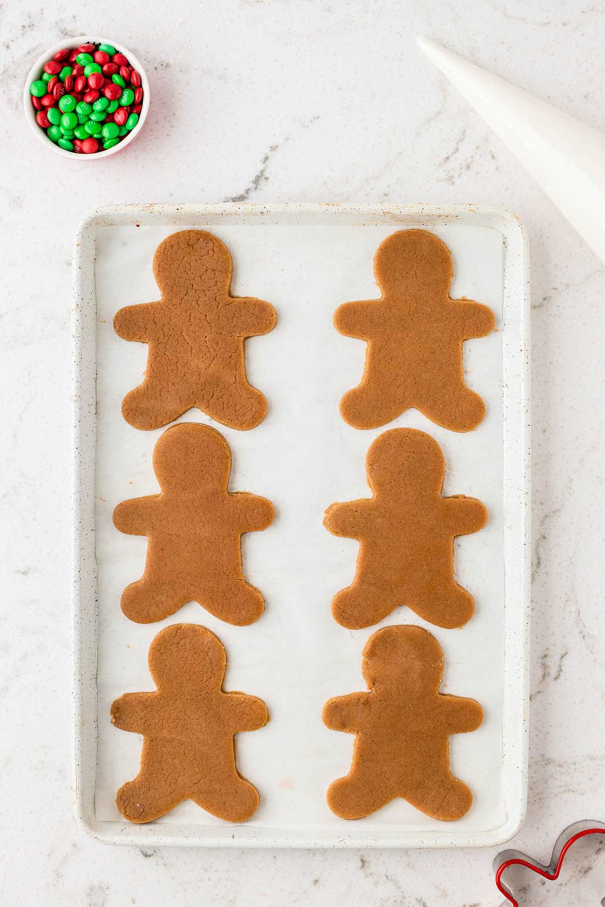 Six undecorated Easy Gingerbread Man Cookies rest on a baking sheet, with a bowl of red and green candies nearby, ready for decorating fun.