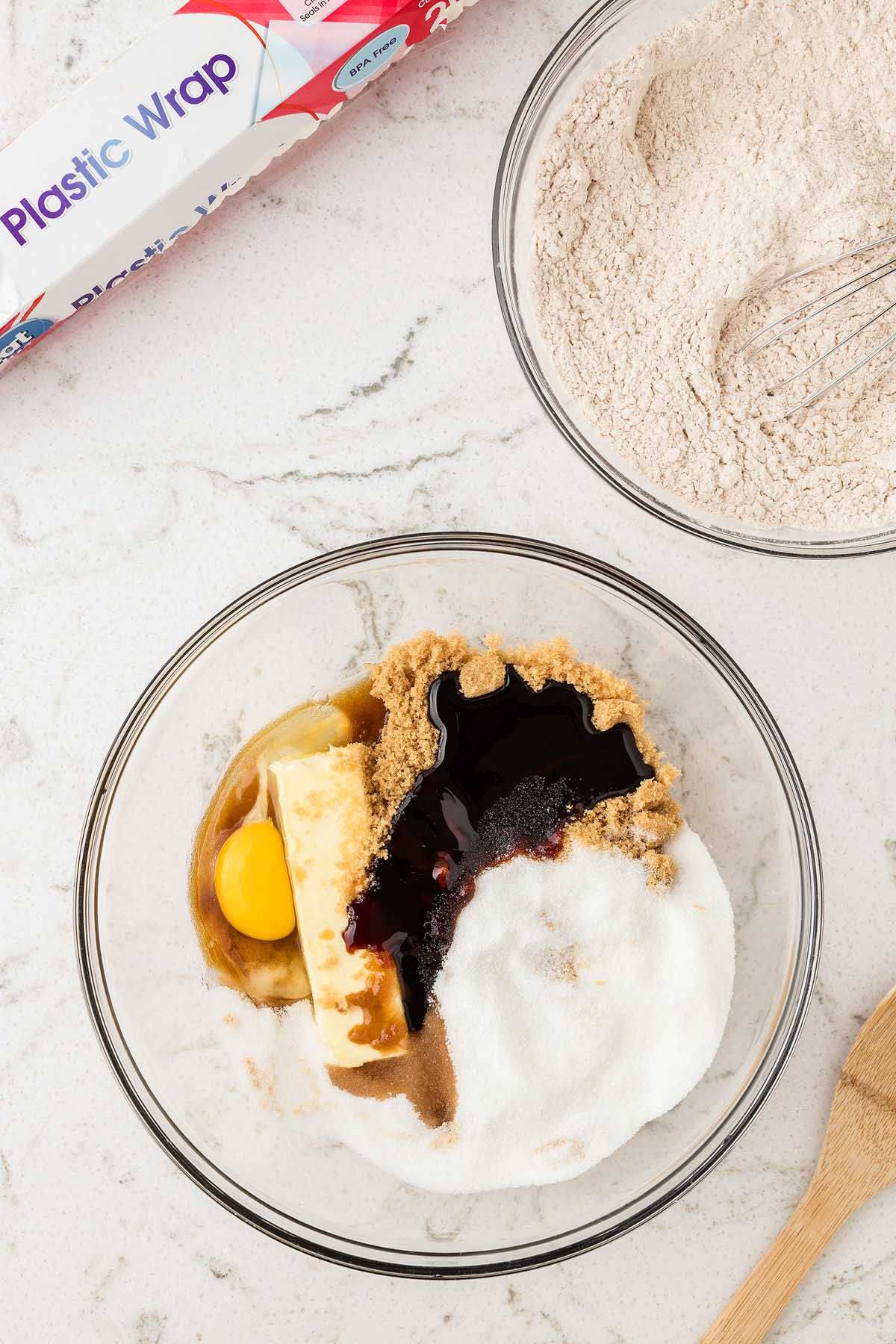 A glass bowl with baking ingredients for Easy Gingerbread Man Cookies, a bowl of flour, plastic wrap, and a wooden spoon on a counter.