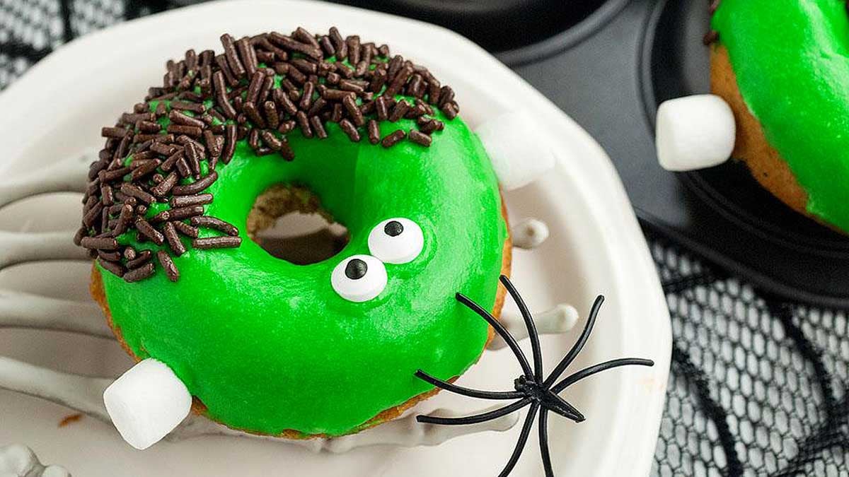 A donut with green icing, candy eyes, chocolate sprinkles, marshmallows, and a toy spider on a plate—perfect for spooky Halloween desserts.