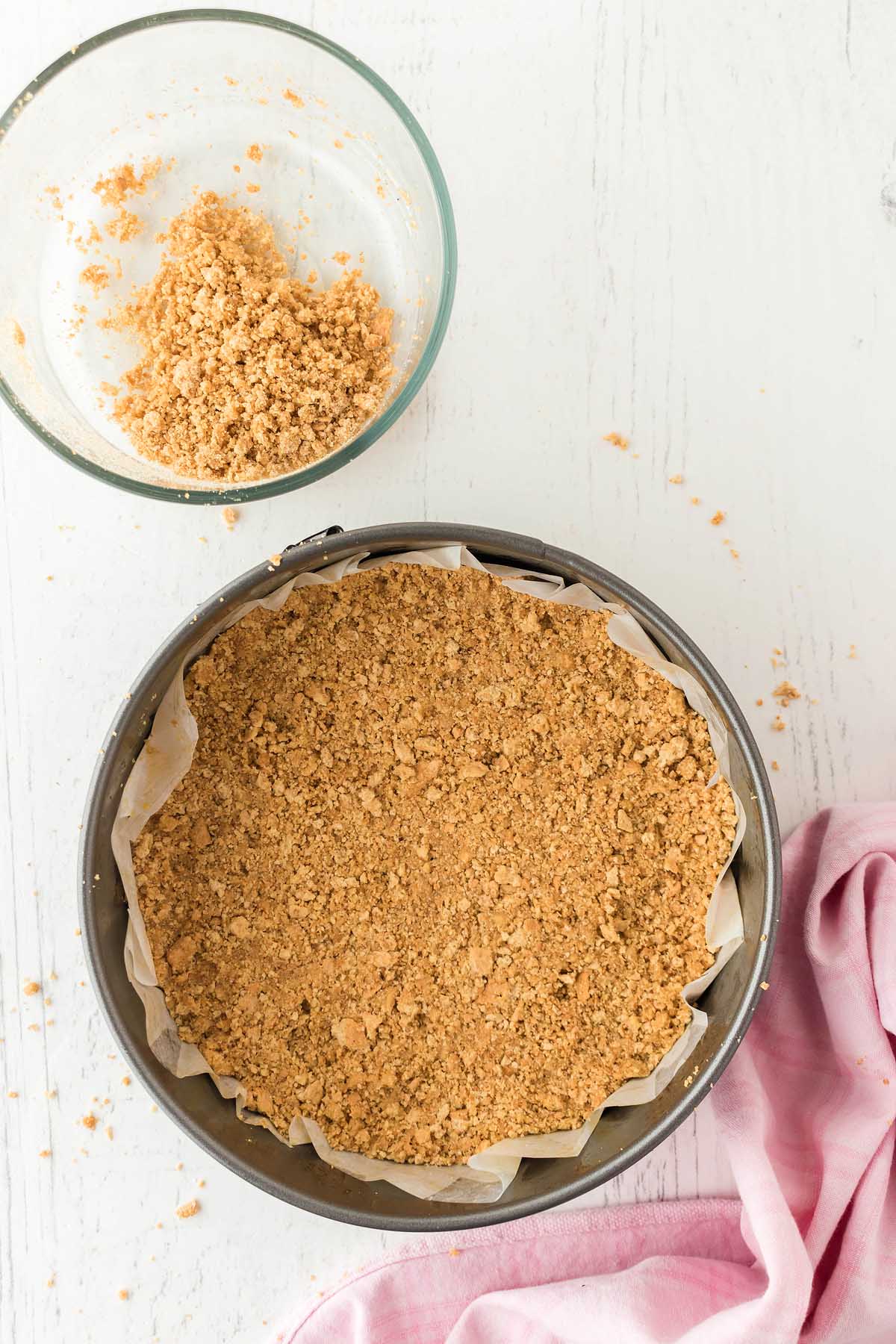 Glass bowl with crumb mixture next to a springform pan filled with funfetti birthday cheesecake crust on a white surface. Pink cloth partially visible.
