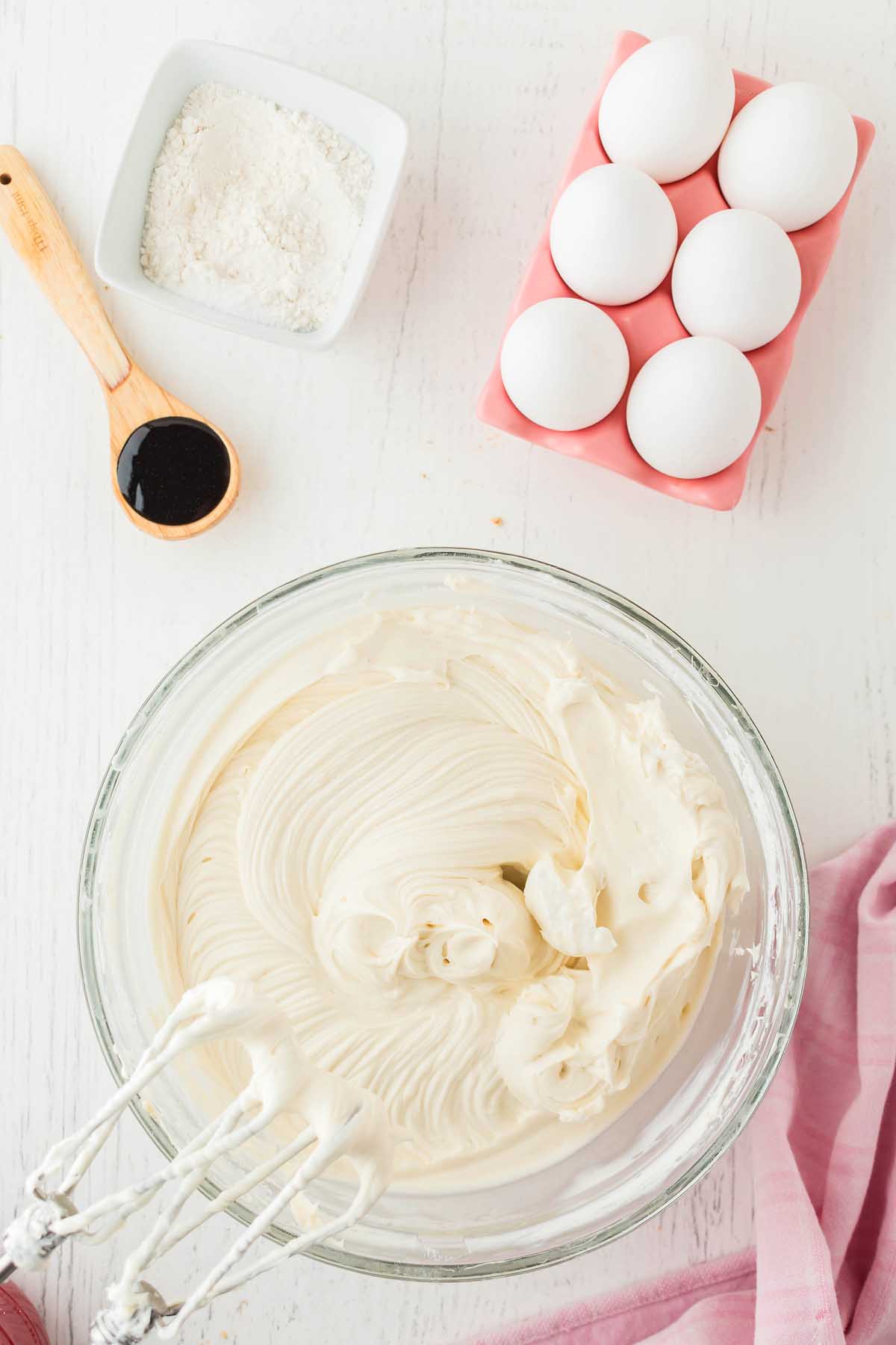 A bowl of creamy batter with a hand mixer hints at the beginnings of a delightful Funfetti Birthday Cheesecake. Nearby, a small dish of flour, a spoon of vanilla extract, and a pink tray holding six eggs sit on a white surface. A pink cloth is partially visible.