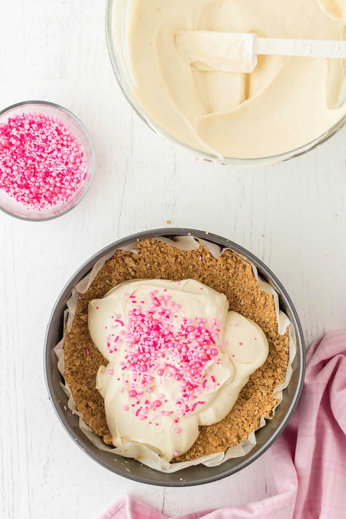 A pie crust in a pan brimming with creamy Funfetti Birthday Cheesecake filling and pink sprinkles. Nearby, a bowl of filling and another of colorful sprinkles sit on a white surface, while a pink cloth is elegantly draped beside the pan.