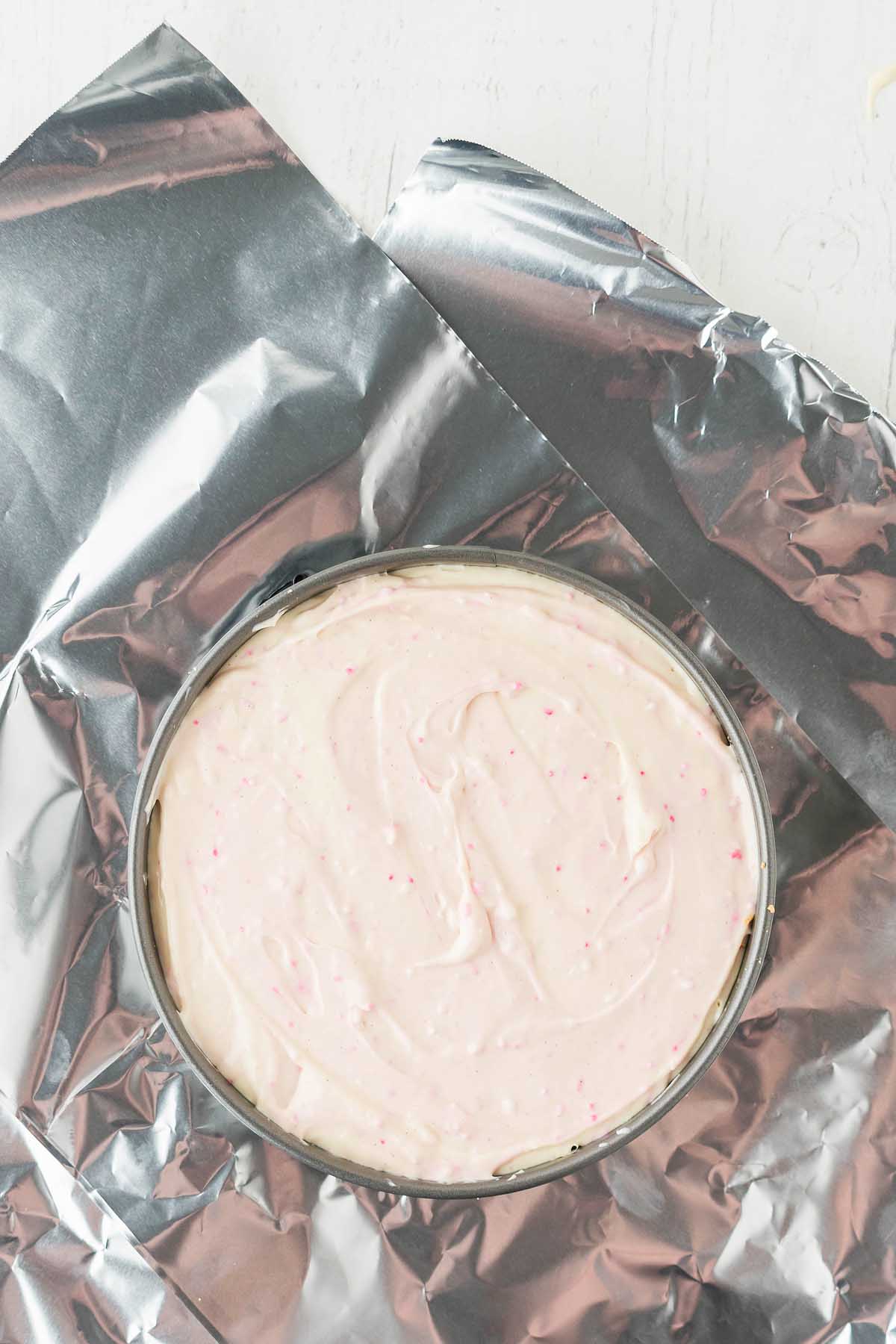A round cake pan filled with light pink Funfetti batter rests on a sheet of aluminum foil, promising a delightful birthday cheesecake.
