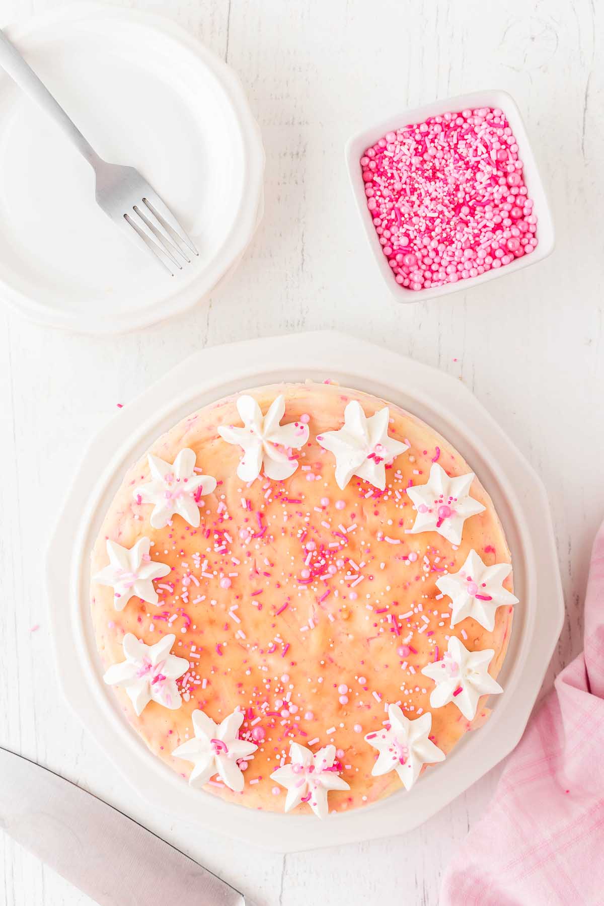 A round Funfetti Birthday Cheesecake with peach-colored icing is topped with white star-shaped meringues and pink sprinkles. Nearby are a fork, a stack of plates, a pink cloth, and a bowl of pink sprinkles.