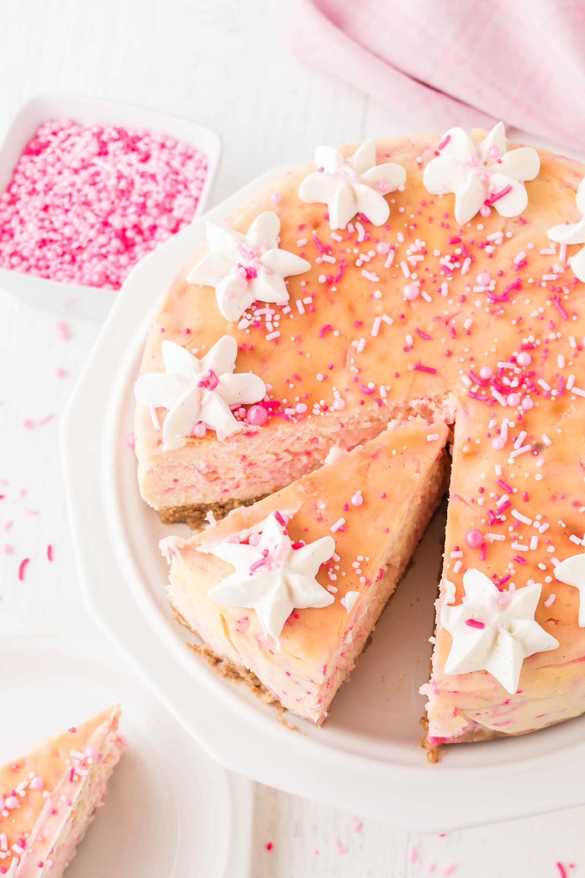 A pink-frosted funfetti cake with white star-shaped whipped cream decorations and pink sprinkles, with one slice cut out. A bowl of pink sprinkles is in the background, adding a playful touch to this delightful birthday cheesecake.
