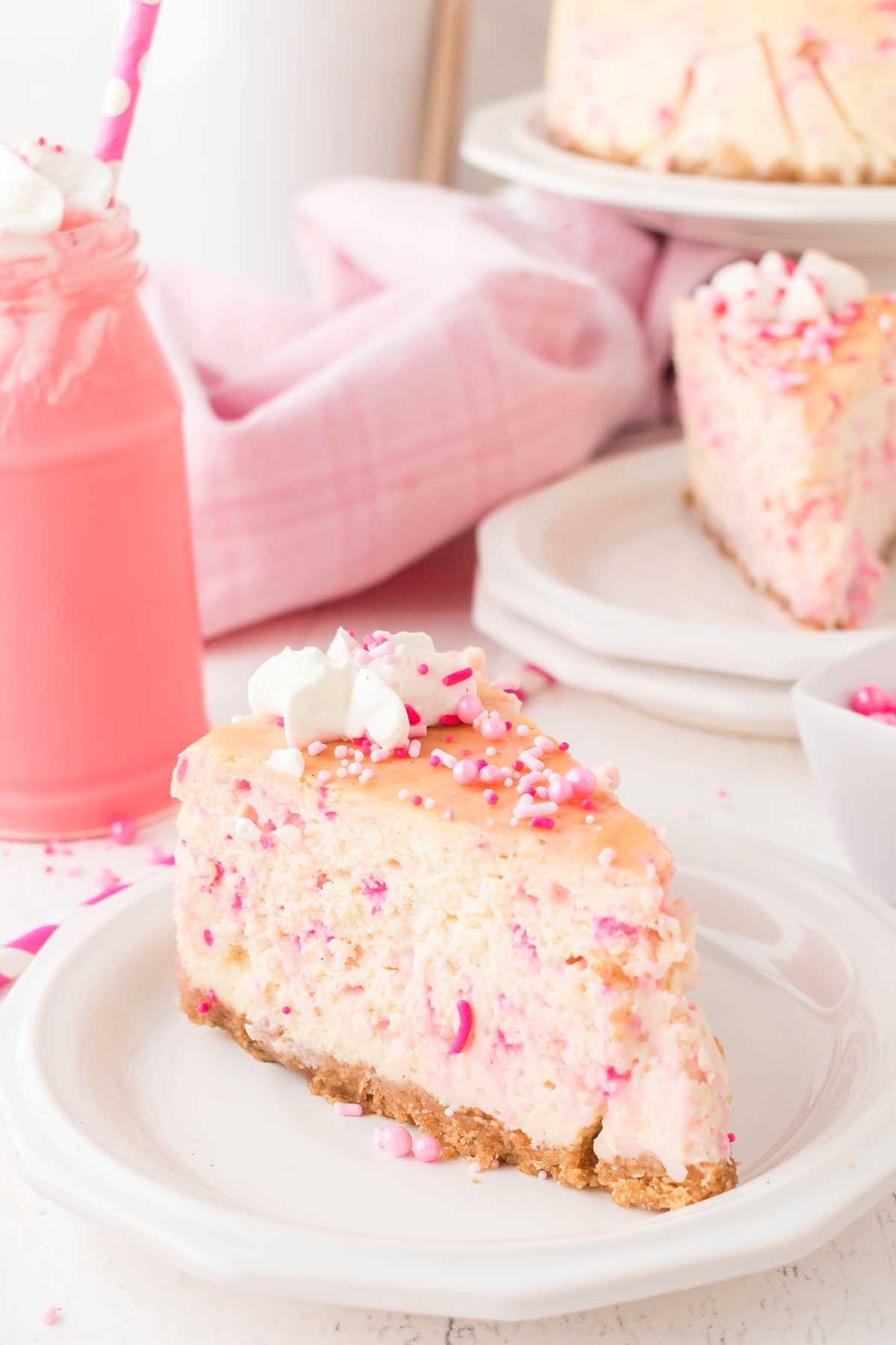 A slice of funfetti birthday cheesecake, complete with colorful sprinkles, sits invitingly on a white plate. Next to it, a pink drink in a glass jar with a straw adds charm. In the background, more slices and a cloth add to the festive scene.