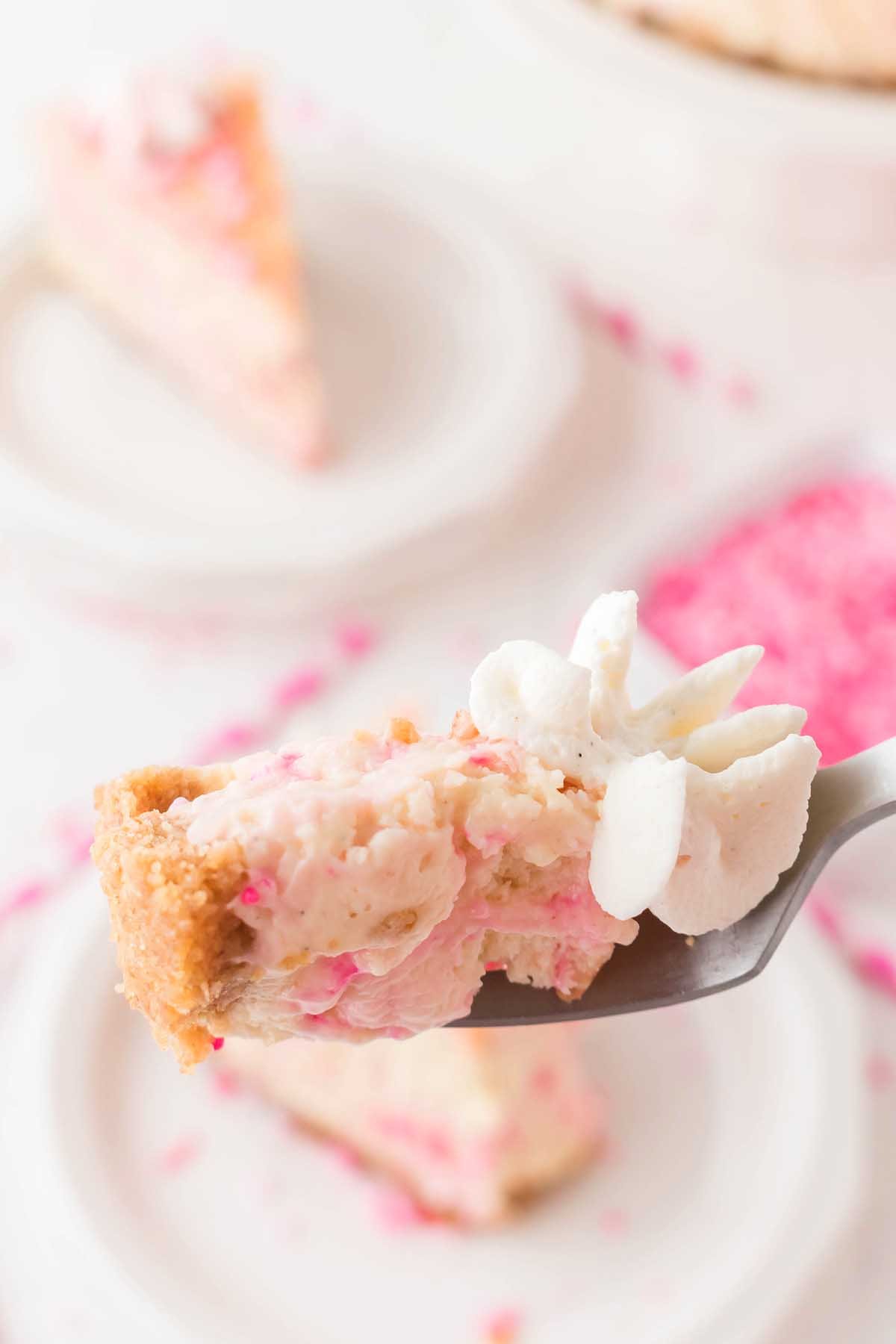 A fork holds a bite of Funfetti Birthday Cheesecake with a graham cracker crust and whipped cream, sprinkled with colorful confetti pieces. More slices are artfully blurred in the background, adding to the festive delight.