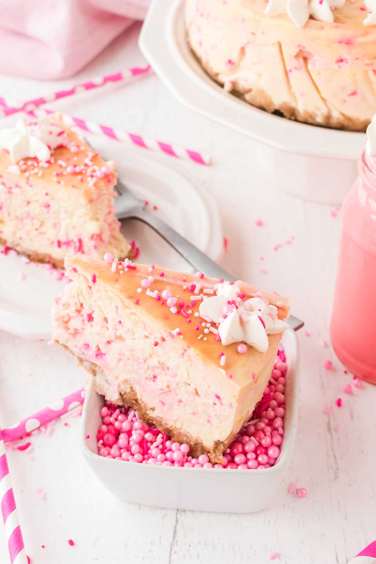 A slice of Funfetti Birthday Cheesecake with whipped cream and sprinkles sits on a small dish. In the background, other slices and a pink drink set the celebratory scene.