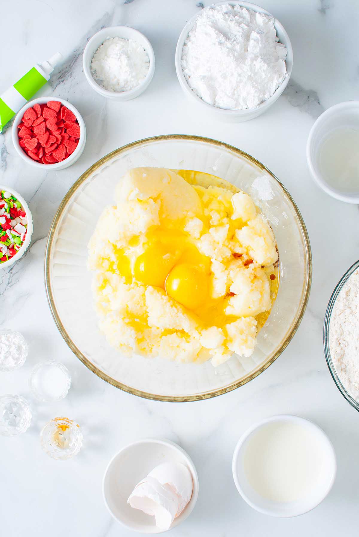 A bowl with eggs, sugar, and butter, surrounded by flour, sprinkles, and baking ingredients on a white counter—perfect for whipping up festive Grinch Crumbl Christmas Sugar Cookies.