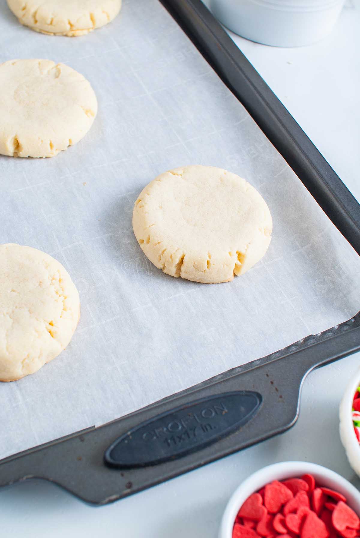 Unbaked Grinch Crumbl Christmas Sugar Cookies on a parchment-lined baking sheet, with bowls of colorful sprinkles nearby.