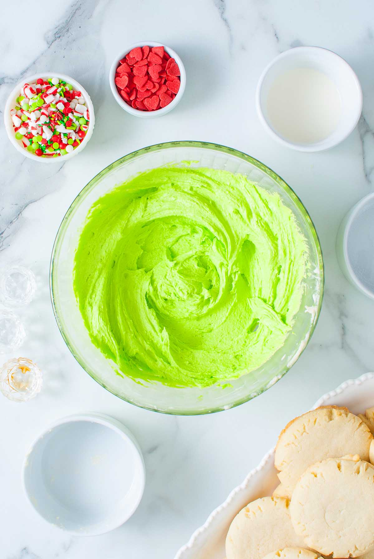 Large bowl of bright green frosting, perfect for decorating Grinch Crumbl Christmas Sugar Cookies, surrounded by cookies, sprinkles, and small bowls on a marble surface.