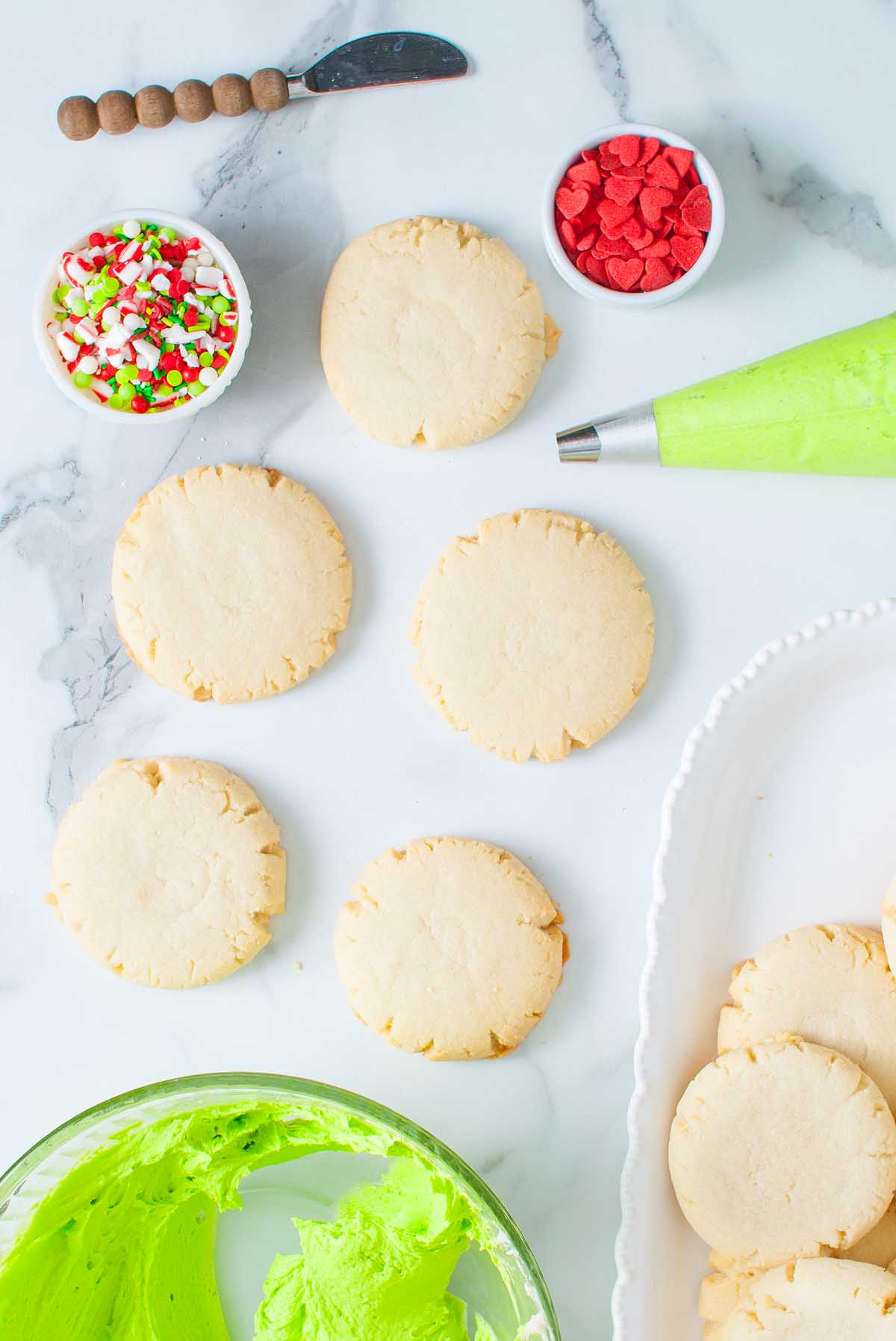 Round sugar cookies inspired by Grinch Crumbl Christmas Sugar Cookies, bowls of colorful sprinkles, red candies, and green frosting on a marble surface.