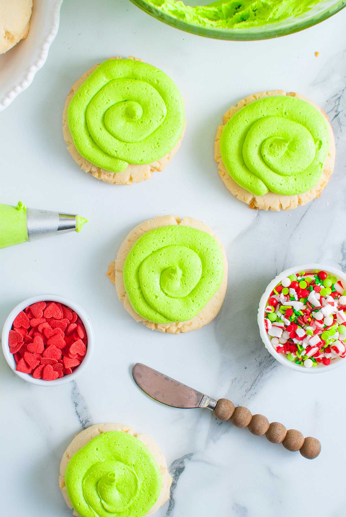 Four Grinch Crumbl Christmas Sugar Cookies with green frosting, surrounded by red and holiday sprinkles, knife, and piping bag.
