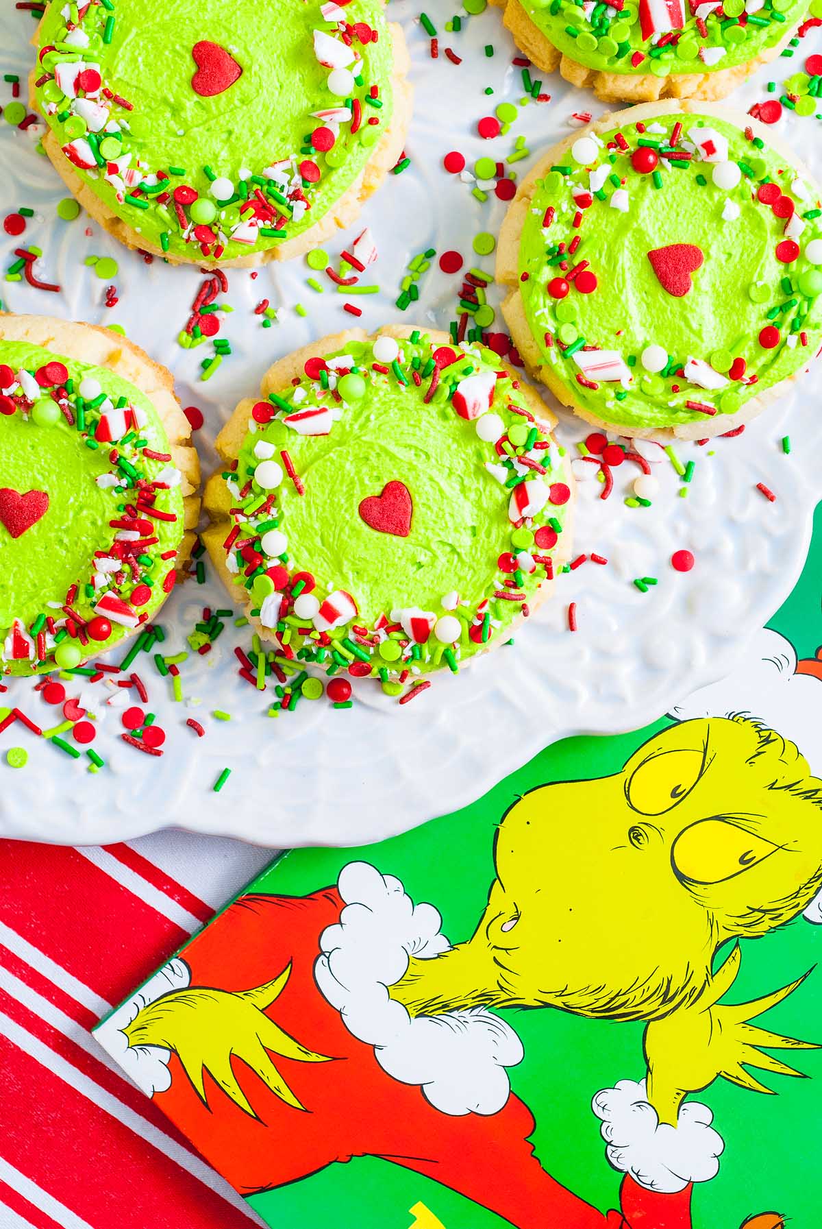Green-frosted Grinch Crumbl Christmas Sugar Cookies with sprinkles and heart decor sit beside a classic Dr. Seuss Grinch book.