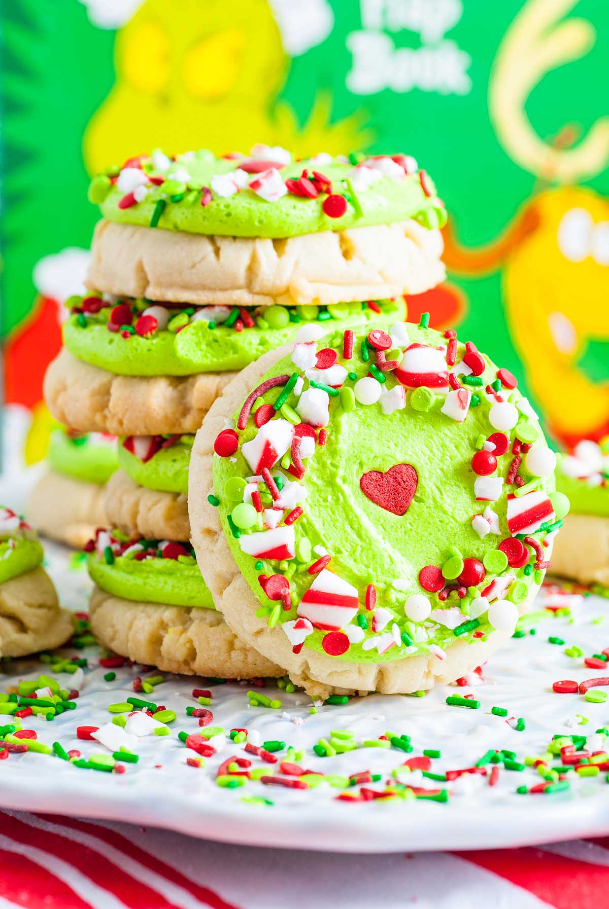 Stack of frosted Grinch Crumbl Christmas Sugar Cookies with green icing, red heart, and festive sprinkles on a white plate.