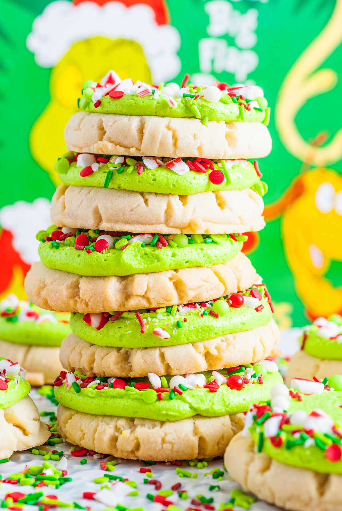A stack of festive Grinch Crumbl Christmas Sugar Cookies with green frosting and colorful holiday sprinkles sits in front of a playful Grinch backdrop.