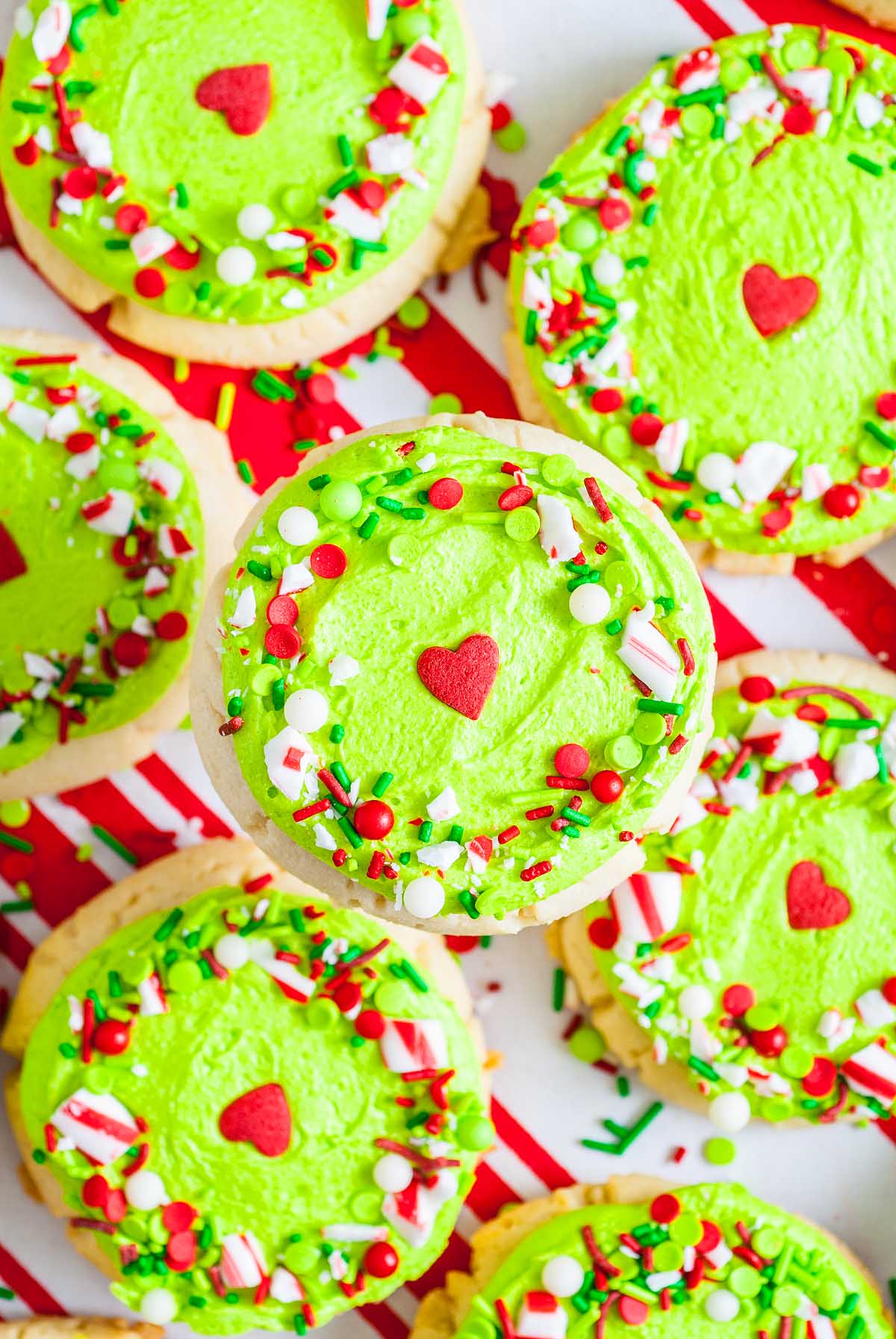 Round Grinch Crumbl Christmas Sugar Cookies topped with bright green frosting, sprinkles, and a red heart candy in the center are displayed on a festive striped background.
